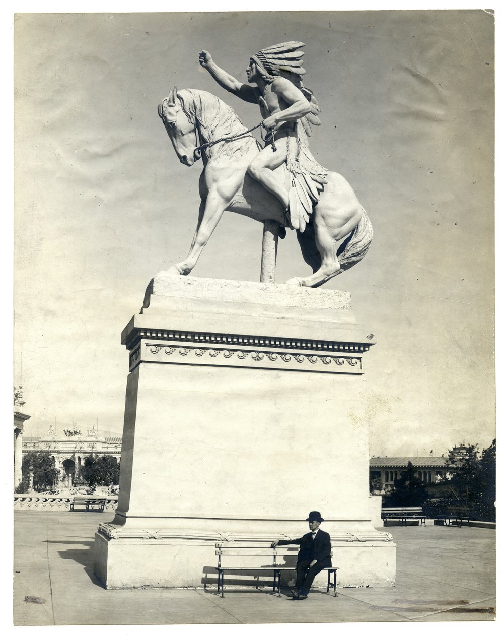 Vertikales, sepiafarbenes Foto einer Statue eines amerikanischen Indianers auf einem Pferd auf einem großen Sockel. Ein Mann im dunklen Anzug und Hut sitzt auf einer Bank am Sockel von Unknown