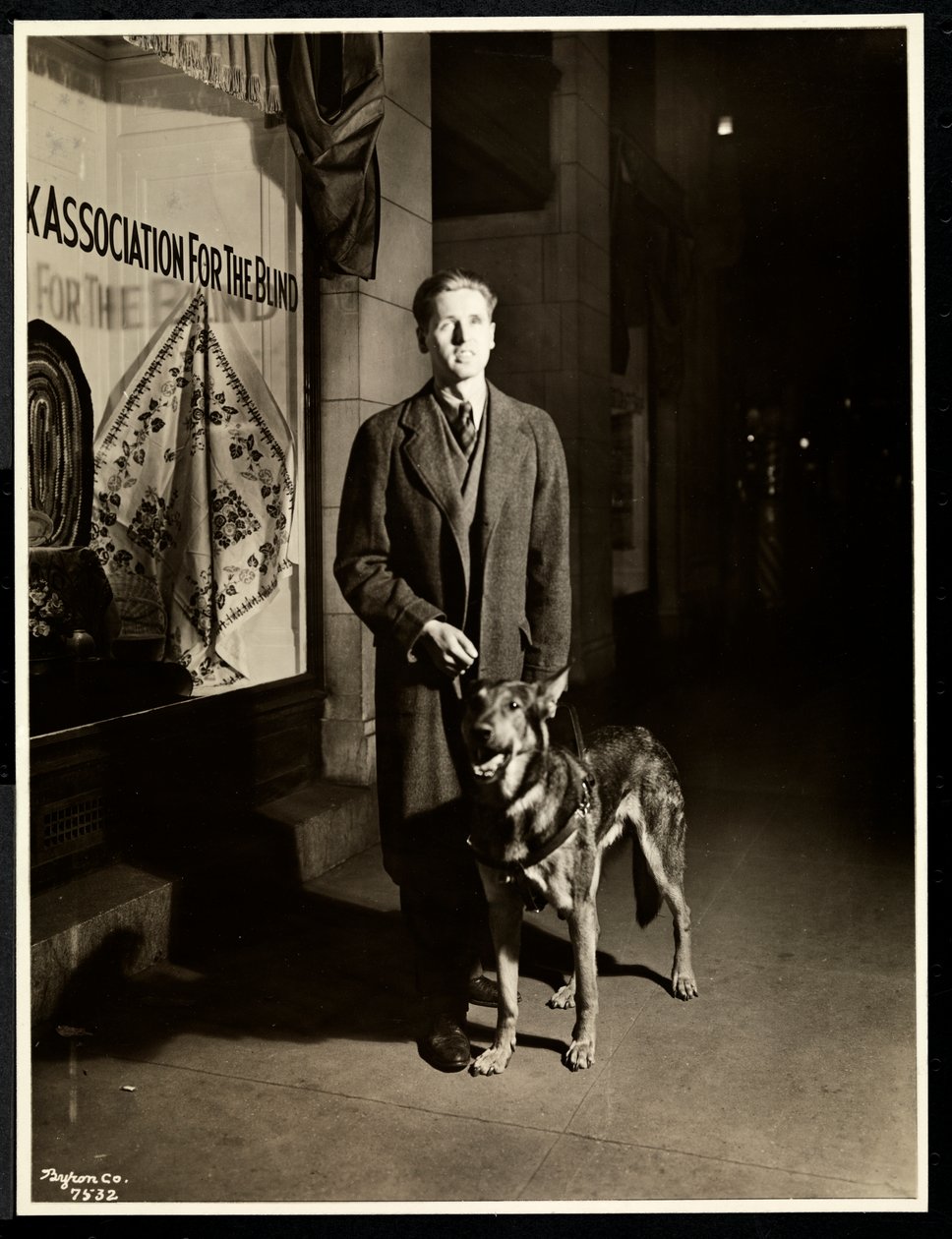 Blinder Mann und sein Blindenhund auf dem Bürgersteig vor der New York Association for the Blind, 111 East 59th Street, New York, 1938 (Silbergelatineabzug) von Byron Company