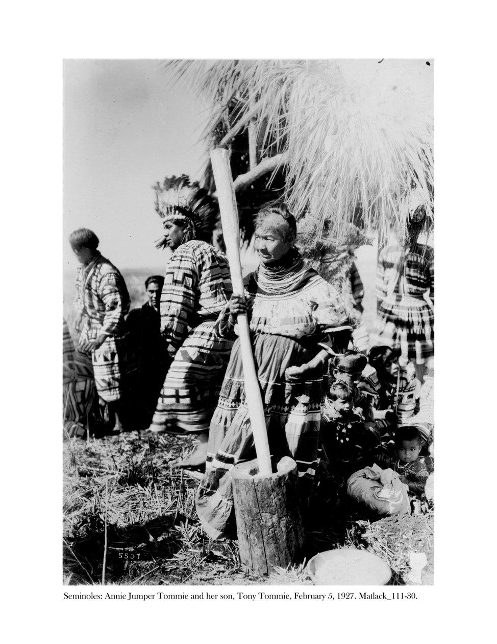 Annie Jumper Tommie benutzt einen Mörser und Stößel, um Maismehl herzustellen, und ihr Sohn, Tony Tommie, trägt einen Adlerfederkopfschmuck, 5. Februar 1927 von Claude C. Matlack