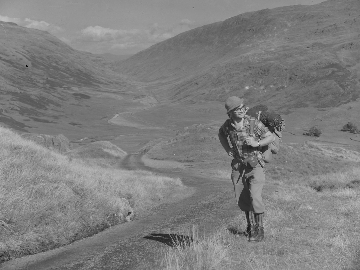 Ein Mann geht mit einem Rucksack auf dem Rücken in einer offenen Tal-Landschaft von Joseph Hardman