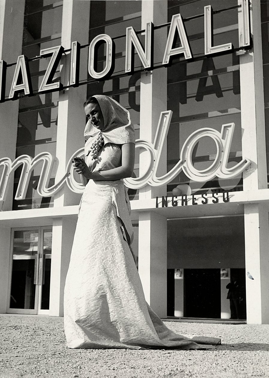 Modell im Hochzeitskleid des Modehauses "Lamma". Der Artikel zeichnet sich durch einen Kopfschmuck im klösterlichen Stil aus, der am Hals mit einer auffälligen Traubenbrosche gehalten wird. von Luis Bogino