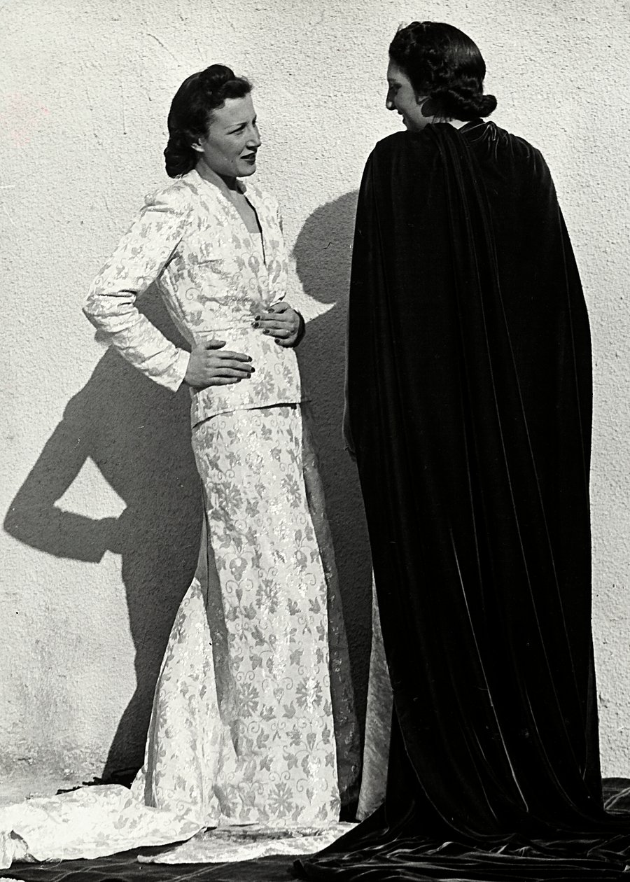 Zwei Modelle präsentieren Kreationen des italienischen Modehauses "Dragoni". Ein Modell trägt ein Outfit aus gemustertem Material, bestehend aus einer Jacke und einem langen Rock mit Schleppe. Das andere Modell, von hinten gesehen, trägt einen langen Samtu von Luis Bogino