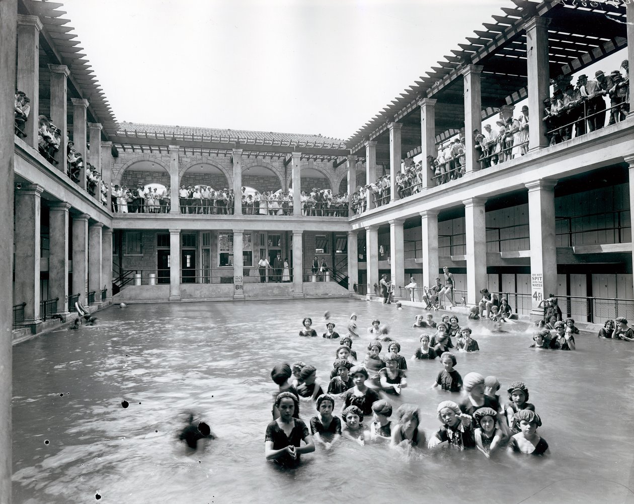Horizontales Schwarz-Weiß-Foto eines Schwimmbeckens mit zweistöckigem Balkon, der von Säulen gestützt wird. Zuschauer beobachten die Schwimmer vom zweiten Balkon von William G. Swekosky