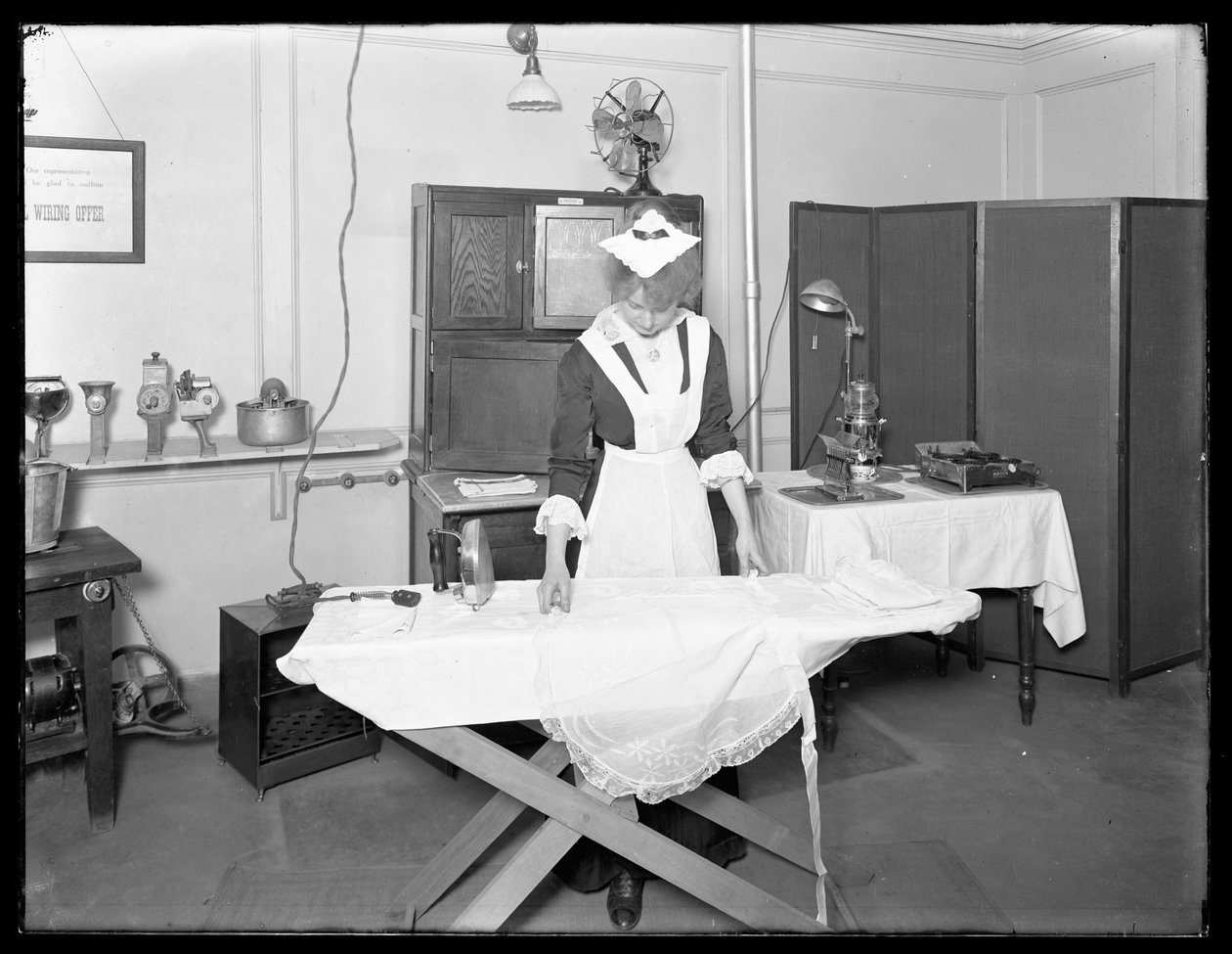 Junge Frau in einem Dienstmädchenkostüm demonstriert die Verwendung eines elektrischen Bügeleisens, ca. 1913-14 von William Davis Hassler