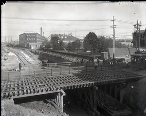 Horizontales Schwarz-Weiß-Foto einer Holzbrücke und Straßenbahnschienen über teilweise gebaute Eisenbahnschienen. Eine Straßenbahn mit der Aufschrift Chouteau 2137 beginnt rechts die Brücke zu überqueren. Im Hintergrund Fabrikgebäude mit der Aufschrift Sta von Charles Clement Holt