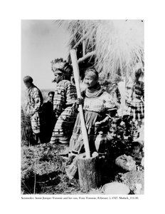 Annie Jumper Tommie benutzt einen Mörser und Stößel, um Maismehl herzustellen, und ihr Sohn, Tony Tommie, trägt einen Adlerfederkopfschmuck, 5. Februar 1927