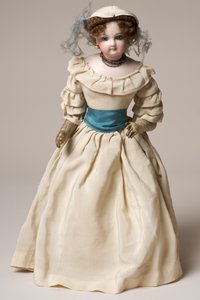 Biskuitmodepuppe mit schwenkbarem Kopf, trägt ein langes cremefarbenes Kleid und einen Hut