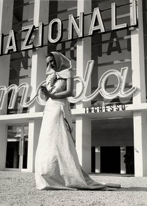 Modell im Hochzeitskleid des Modehauses "Lamma". Der Artikel zeichnet sich durch einen Kopfschmuck im klösterlichen Stil aus, der am Hals mit einer auffälligen Traubenbrosche gehalten wird. von Luis Bogino