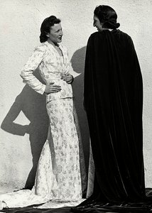 Zwei Modelle präsentieren Kreationen des italienischen Modehauses "Dragoni". Ein Modell trägt ein Outfit aus gemustertem Material, bestehend aus einer Jacke und einem langen Rock mit Schleppe. Das andere Modell, von hinten gesehen, trägt einen langen Samtu von Luis Bogino
