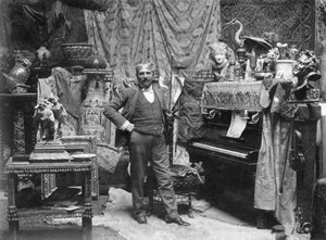 Ganzkörperporträt des Malers Francesco Vinea im Atelier von Donatello in Florenz. Er posiert in einem Raum, der mit zahlreichen Vorhängen, chinesischen Vasen, kleinen Skulpturen und Nippes dekoriert ist. Rechts ein Klavier und eine Geige (s/w Foto) von Mario Nunes Vais