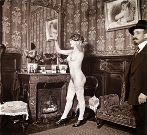 Rätselhaftes Foto, das eine junge Frau völlig nackt (außer Schuhe und Strümpfe) zeigt, die sich vor einem Spiegel in Anwesenheit eines Mannes, der sich kleidet und einen Hut trägt, die Haare richtet. Man könnte denken, es sei ein Foto in einem Bordell, abe von Unbekannter Künstler