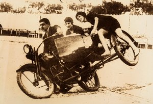 Ein Mann zeigt sich beim Fahren eines Motorrads mit Beiwagen, der drei junge Frauen trägt. Der Beiwagen hebt sich vom Boden ab, als sie eine Kurve nehmen. von Unknown photographer