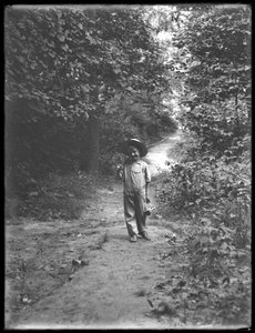 William Gray Hassler mit Strohhut und Latzhose, mit Angelrute und Korb, auf einem Waldweg gehend