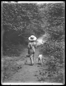 William Gray Hassler mit Strohhut und Latzhose, trägt eine Angelrute und einen Korb, geht einen Weg im Wald entlang mit einem kleinen Hund (Bessie), 20. August 1911 von William Davis Hassler