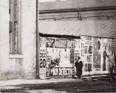 Ein Mann geht an einer Wand mit Plakatwerbung vorbei, darunter eine für Johnnie Hoey, den Jungen Detektiv, der im Havlin
