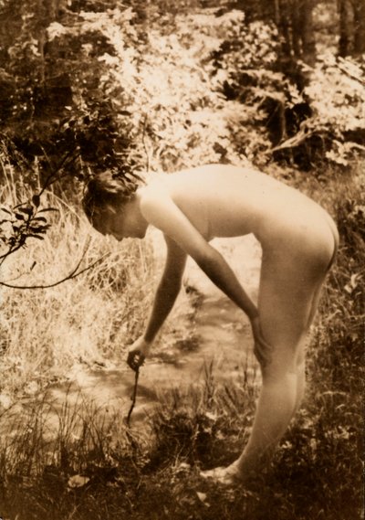 Eine junge Frau, nackt im Wald, im Profil zu sehen, wie sie sich bückt, um einen Zweig in einen Bach zu legen. von Unknown photographer