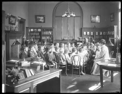 Kinder hören einer Frau im Lesesaal einer nicht identifizierten Filiale der Queens Borough Public Library zu, ca. 1910 von William Davis Hassler