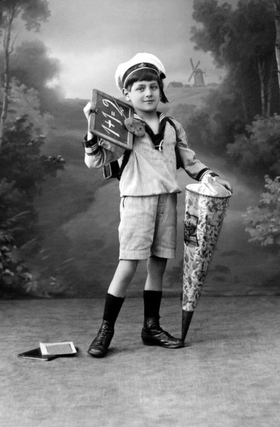 Erster Schultag. Ein Junge in Uniform posiert mit einer Schiefertafel, die die Addition 1+1 = 2 zeigt, und einer Überraschungstasche. Fotografie um 1910. von Ferdinand Georg Waldmuller