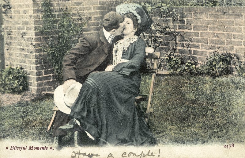 Mann stiehlt einen Kuss von einem Mädchen auf einer Gartenbank von English Photographer