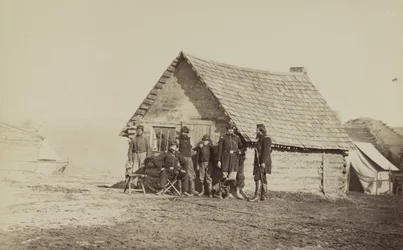 Eine Gruppe von Soldaten und zwei junge Männer, einer davon ein Afroamerikaner, stehen vor Blockhüttenquartieren, 1861-65