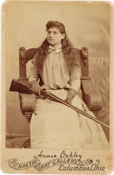 Annie Oakley: Hier sitzt die legendäre Scharfschützin eher schüchtern in einem massiven Renaissance-Stil Stuhl, ein Gewehr ruht auf ihrem Schoß. Ihre zarten weiblichen Züge und fließenden Haare täuschen über ihr robustes Outdoor-Image hinweg