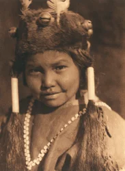 Nahaufnahme eines amerikanischen Indianerjungen vom Stamm oder Dorf der Klamath. Er trägt eine Pelzmütze mit aufgesetzten Eicheln und Federn sowie sein Haar in zwei Pferdeschwänzen