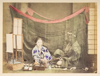 Japanisches Paar in ihrer Freizeit, Studio-Tableau einer jungen japanischen Frau, die mit Teezubehör um sich herum sitzt und eine Pfeife an eine männliche Figur hinter ihr reicht, die von einem Leinen-Moskitonetz abgeschirmt ist, Fotograf: Unbekannt, Lucas