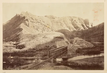 Ansichten der Pazifikbahn, Eisenbahnschienen, die einen Fluss auf einer Fachwerkbrücke überqueren, mit Bergen dahinter