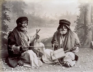 Porträt von zwei arabischen Musikern in bescheidener ethnischer Kleidung. Die älteren Männer posieren im Studio, auf dem Boden sitzend. Hinter ihnen ist ein Hintergrund mit einer bewaldeten Landschaft