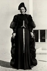 Modell im Abendkleid des italienischen Modehauses "Dragoni". Die Vorderseite des Artikels hat eine auffällige Paillettenverzierung in Form eines Schwertes. Ein Hut und ein Pelzmantel vervollständigen das Ensemble.