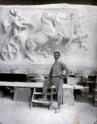 Ganzkörperporträt des berühmten Bildhauers Pietro Canonica. Er posiert in seinem Atelier vor einem Marmorhochrelief, an dem er arbeitet