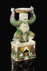 Eiergelb und spinatgrün glasierter Dornleuchter, modelliert als lächelnder Unsterblicher mit geschwungenem Bart und erhobenen Armen, die den auf seinem Kopf balancierten vierpassförmigen Dornaufsatz tragen, Kangxi