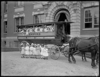 Kleine Mädchen posieren in und vor einem Pferdewagen, vor einem unbekannten Gebäude, wahrscheinlich das Römisch-Katholische Waisenhaus, Kingsbridge, Bronx, ca. 1910