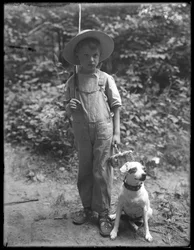 William Gray Hassler mit Strohhut und Latzhose, trägt Angelrute und Korb, posiert im Wald mit einem kleinen Hund (Bessie), 20. August 1911